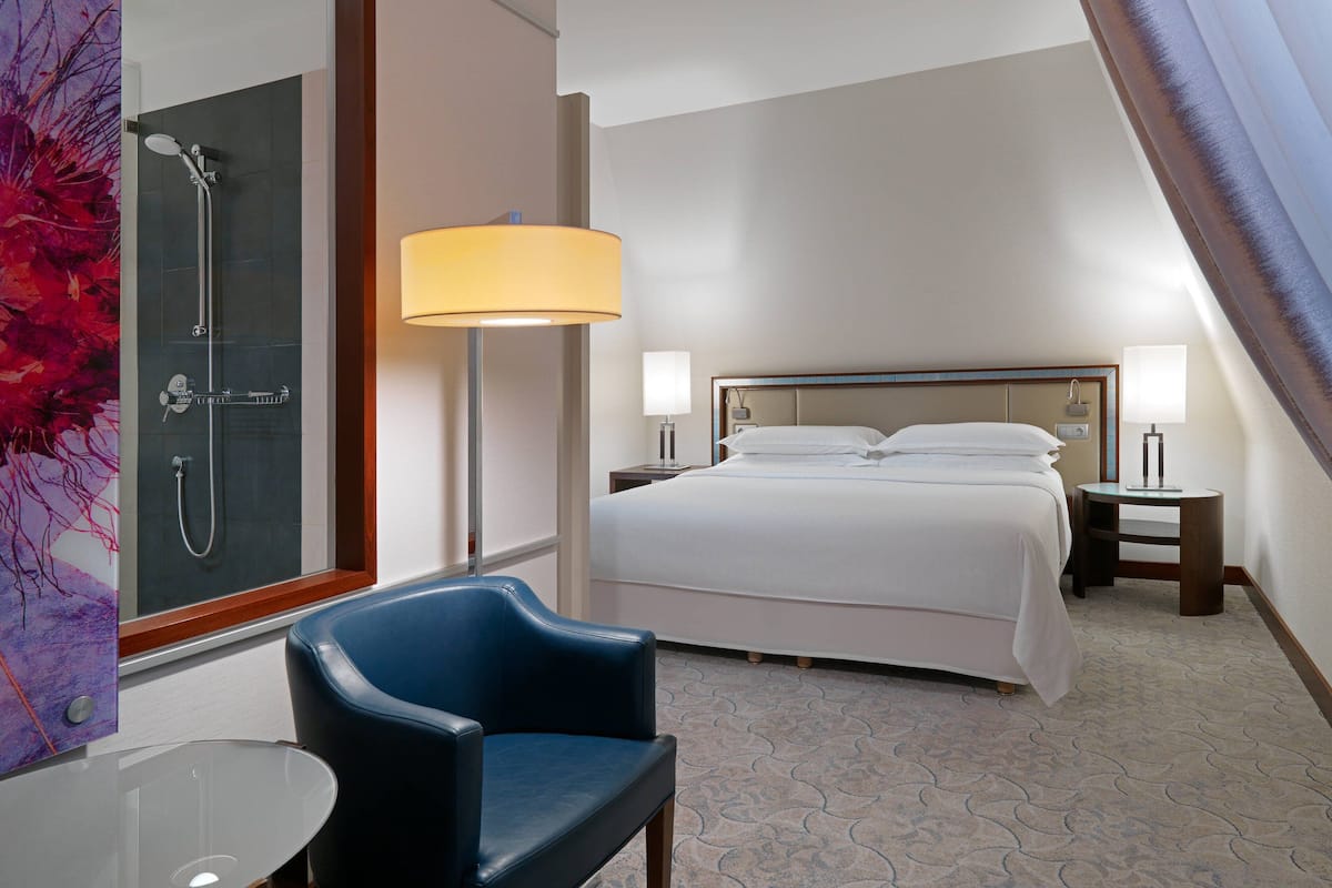 Ein Hotelzimmer mit einem Bett, zwei Nachttischen, einem Sessel, einer Dusche, einer Badewanne mit Glaswand und einer Stehlampe.