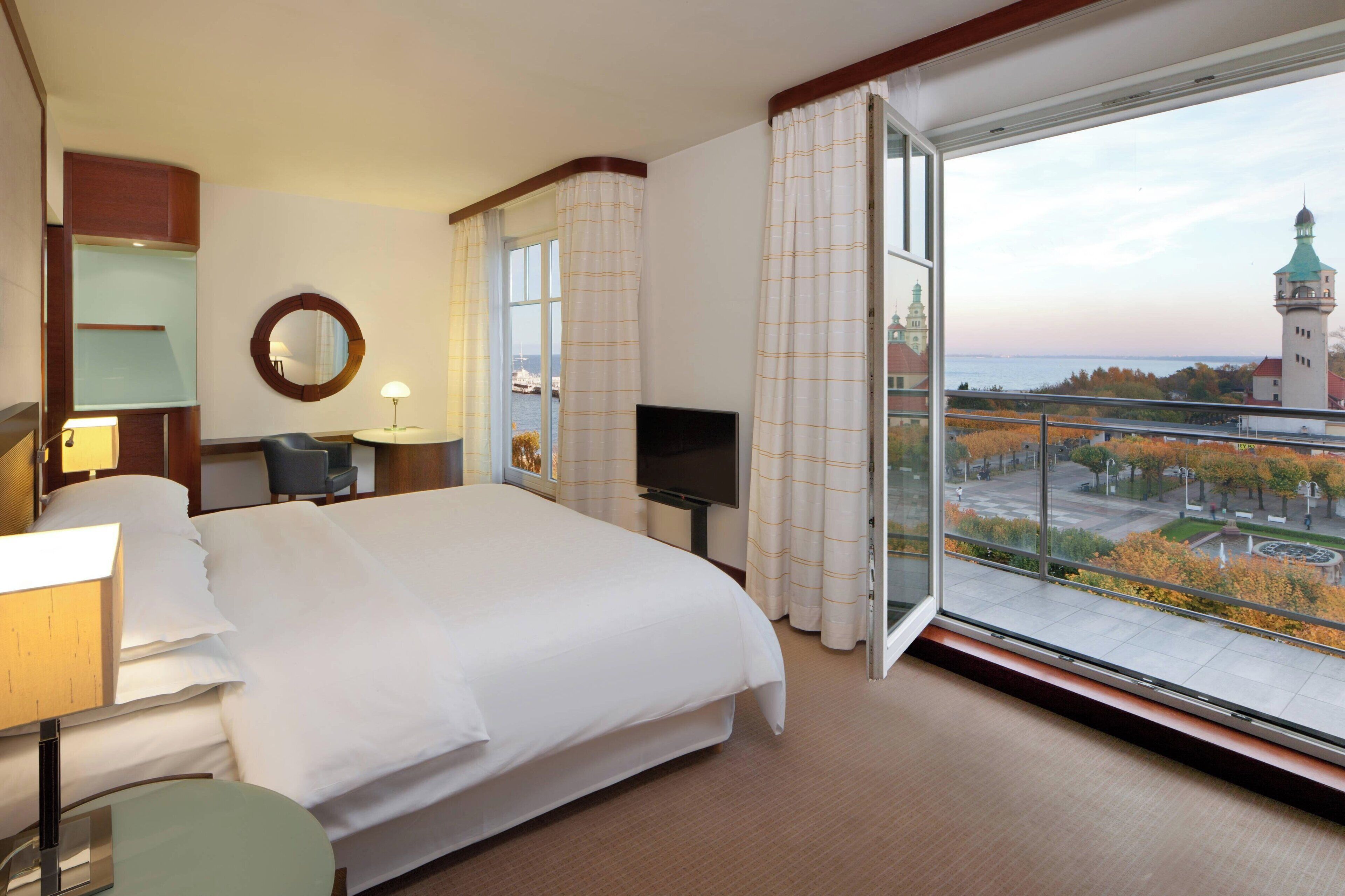 A hotel room with a large bed, a TV, a desk with a lamp, and a view of a tower and trees.
