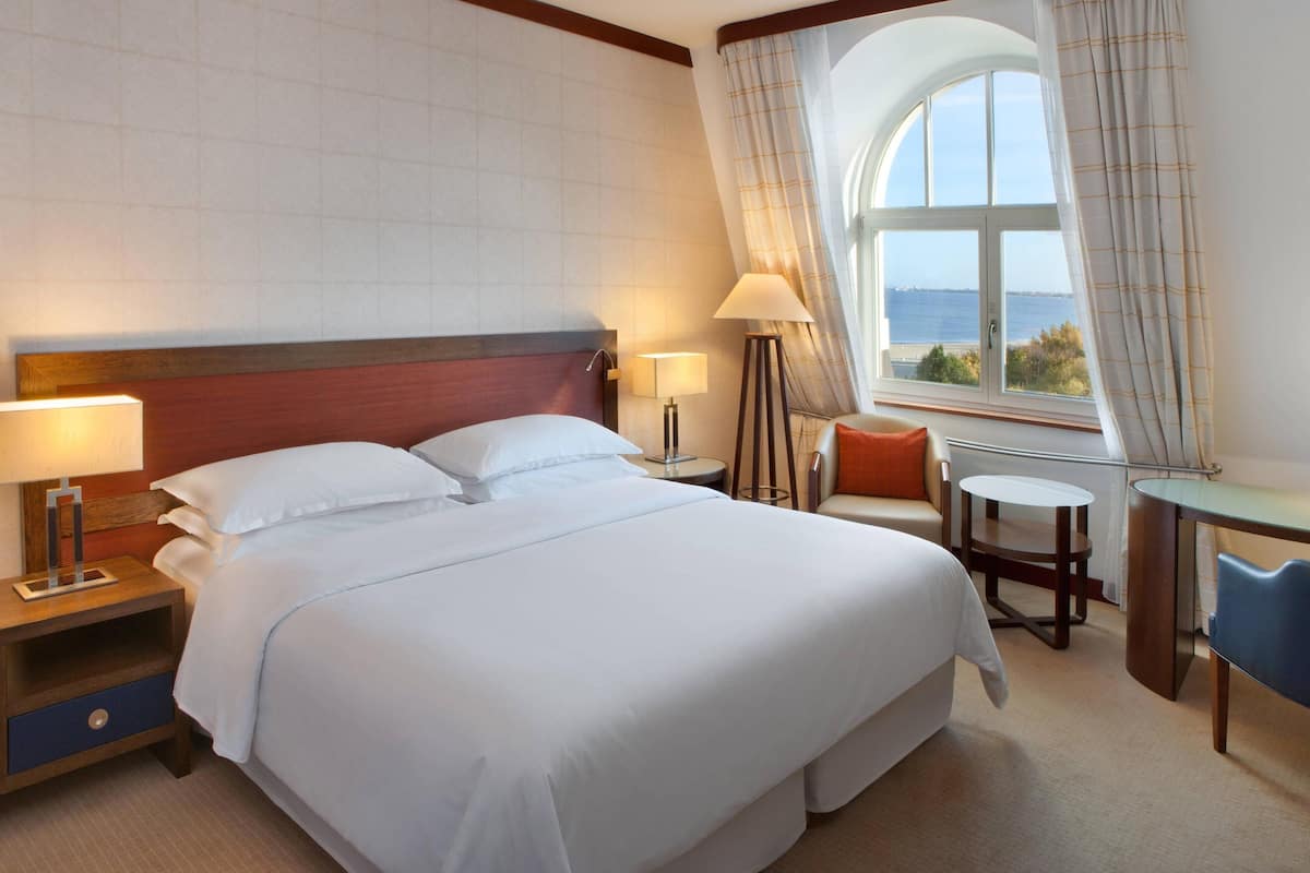 Ein Hotelzimmer mit einem großen Bett, zwei Nachttischen, einem Schreibtisch und Meerblick durch ein Fenster.
