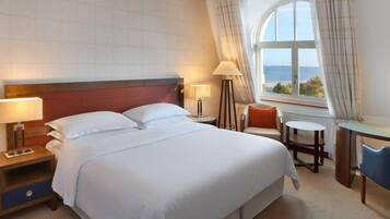 Habitación de hotel con una cama grande, dos mesitas de noche, un escritorio y vistas al mar a través de una ventana.