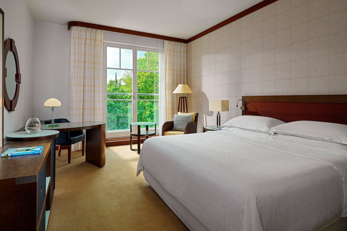 Ein Hotelzimmer mit einem großen Bett, einem Schreibtisch, einem Sessel und einem Fenster mit Vorhängen.