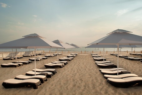 A beach with rows of sun loungers and umbrellas, a sandy path leading to the water, and a clear sky.