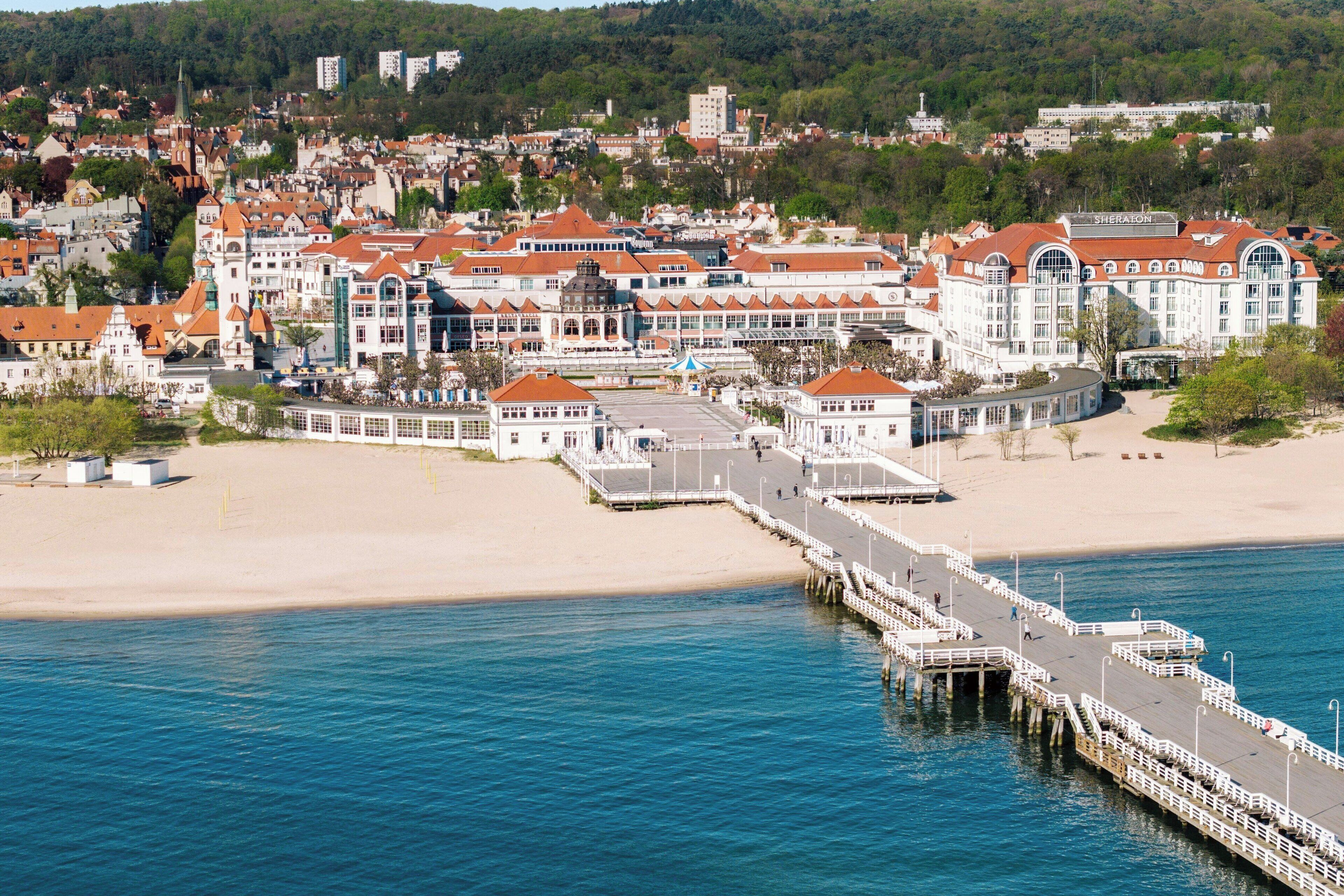 Un pueblo costero con un muelle que se adentra en el mar, rodeado de edificios con techos rojos y una playa de arena.