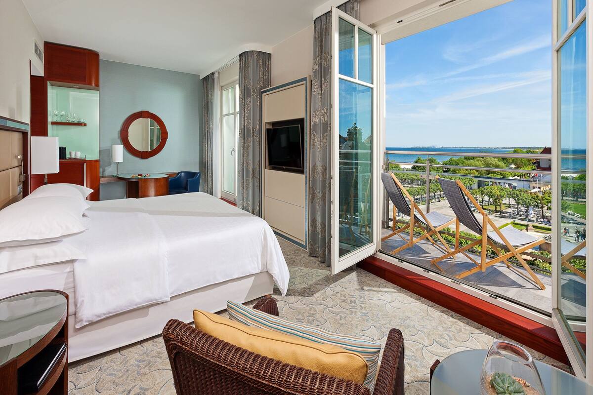Ein Hotelzimmer mit Bett, Fernseher, einem Balkon mit Sitzgelegenheiten und Blick auf die Stadt.