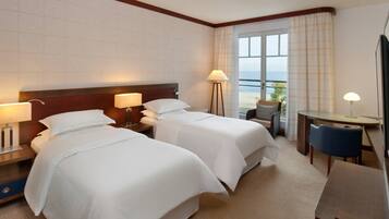 A hotel room with two beds, a desk, a chair, and a view of the sea.