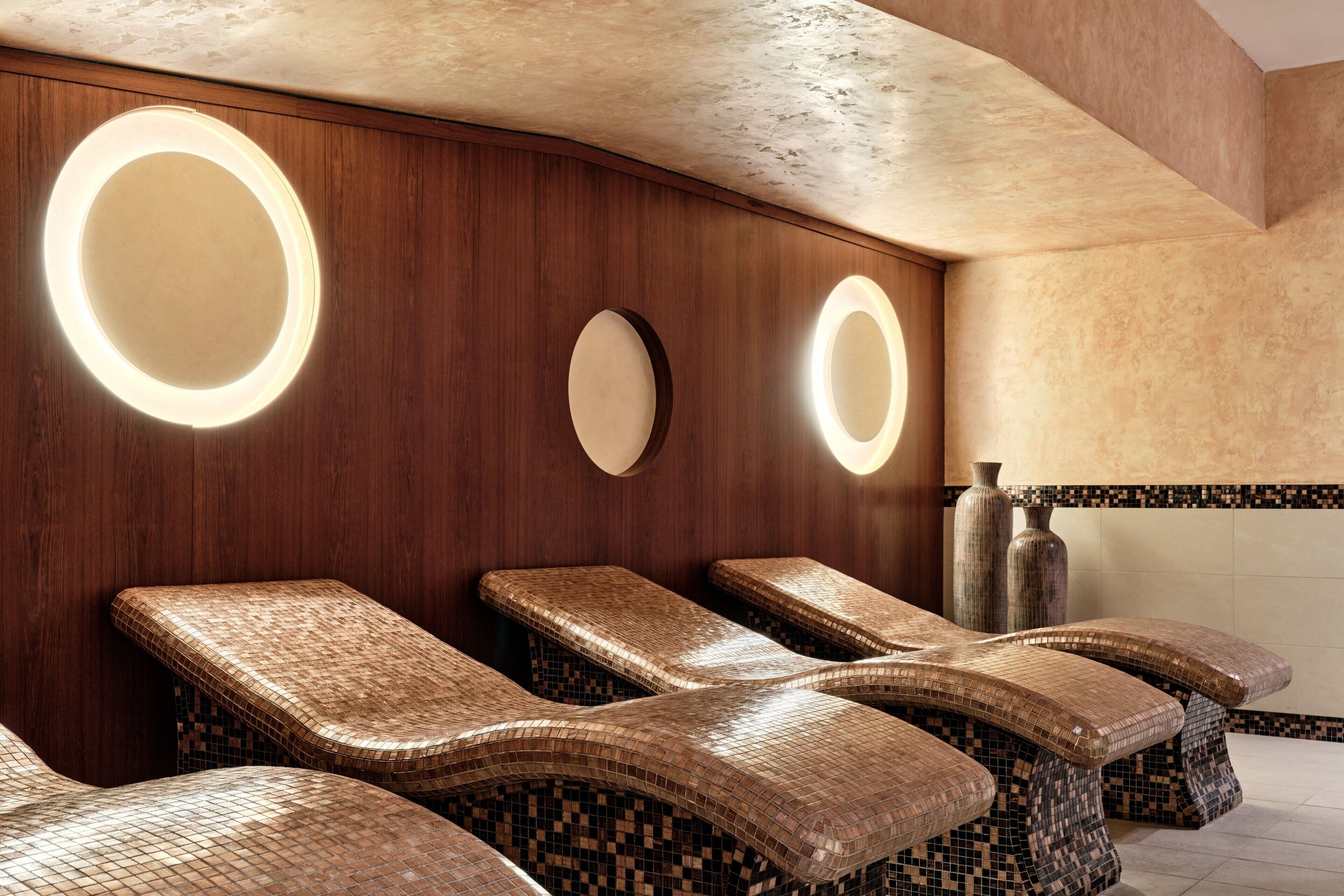 A spa area with mosaic-tiled loungers, wooden walls, and circular mirrors.