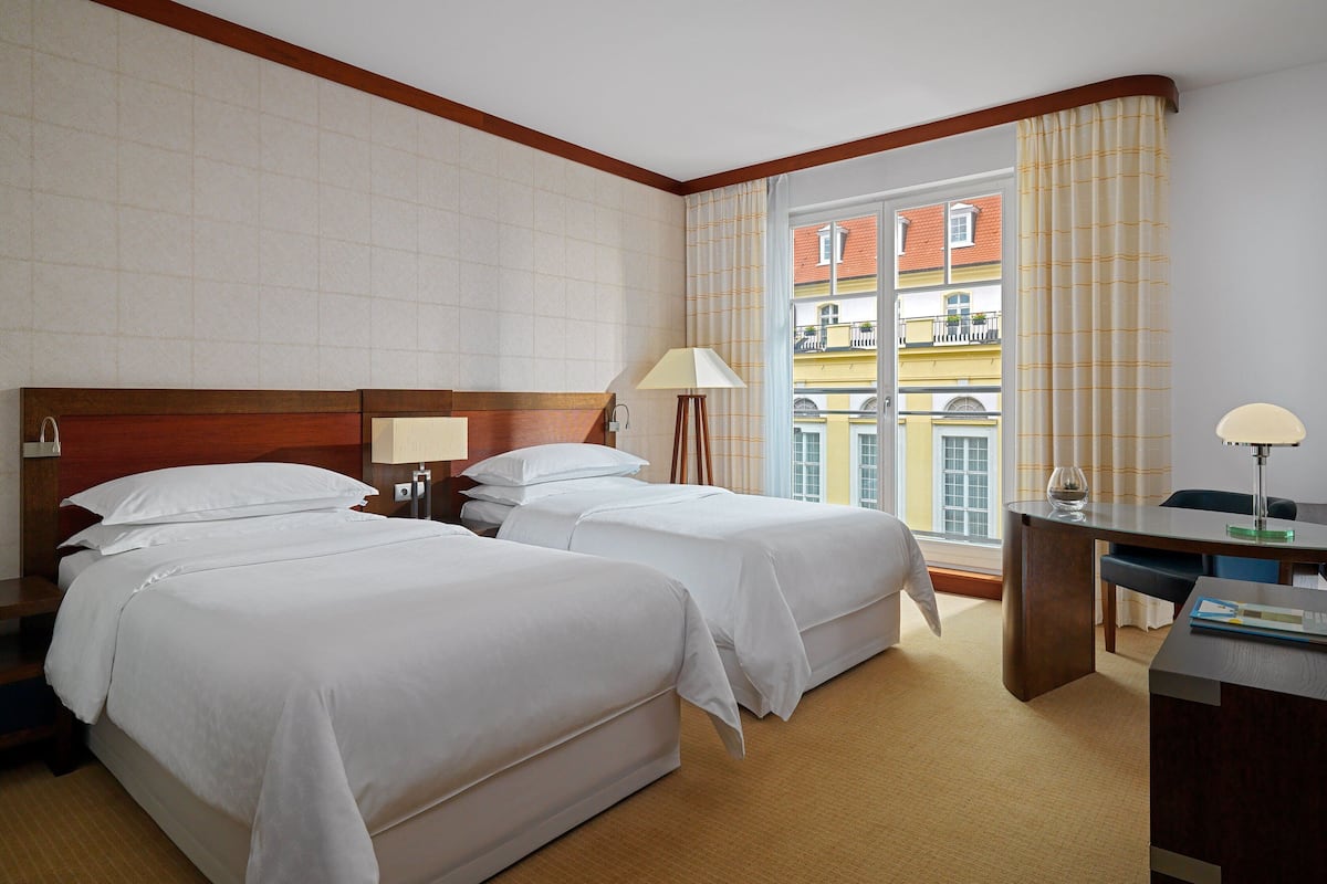 Ein Hotelzimmer mit zwei Betten, einem Schreibtisch und einem Fenster mit Blick auf Gebäude.