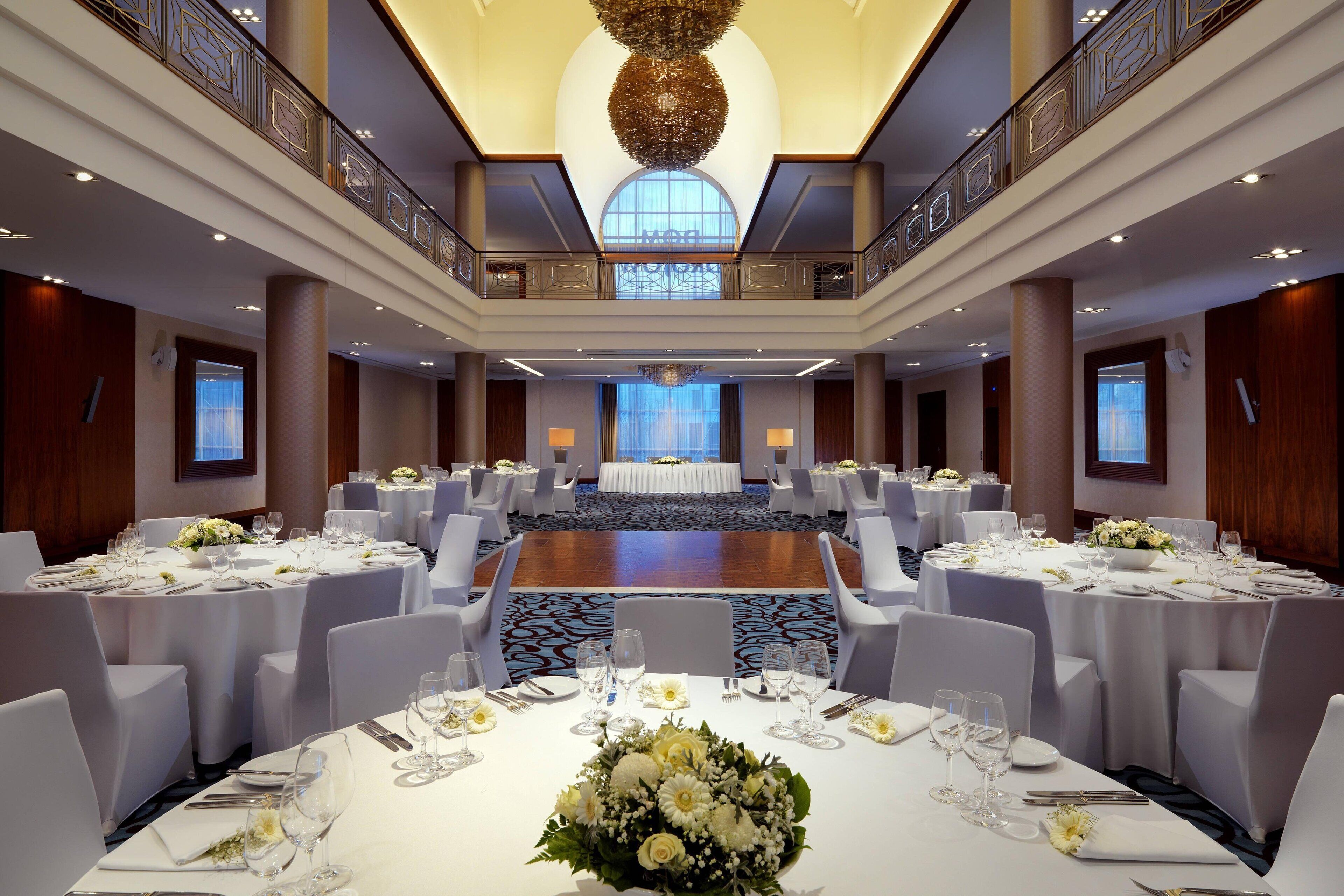 A spacious banquet hall with round tables set for dining, a central stage, and a balcony with decorative railings.