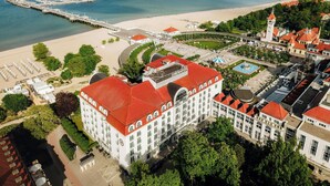 Vista aérea de una zona costera con un edificio grande blanco de techos rojos, un muelle que se adentra en el mar y una playa al fondo.