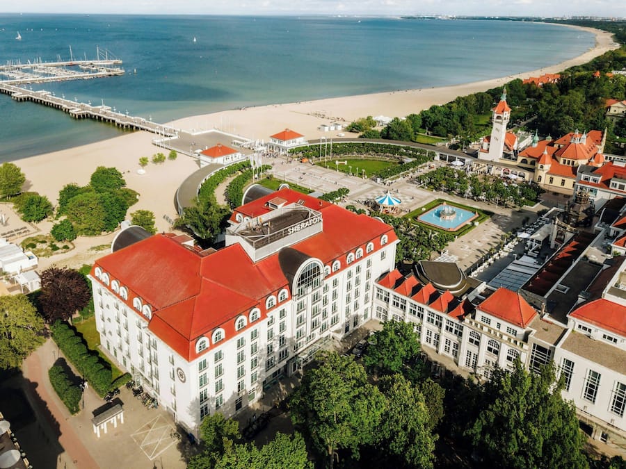 Vue aérienne d’une zone côtière avec un grand bâtiment blanc aux toits rouges, une jetée s’avançant dans la mer et une plage en arrière-plan.