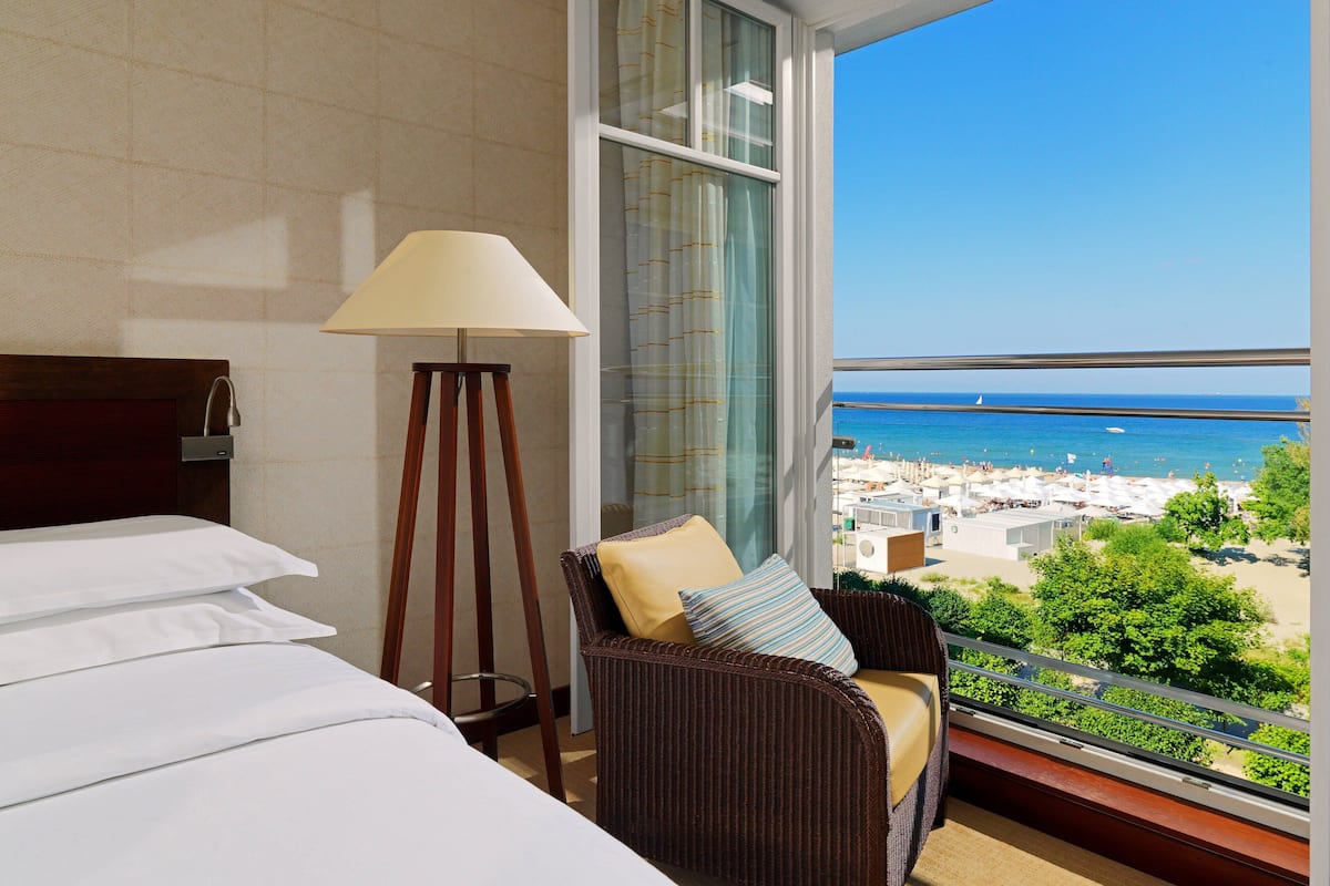 Ein Hotelzimmer mit einem Bett, einem Sessel mit Kissen, einer Stehlampe und einem großen Fenster mit Blick auf den Strand und das Meer.