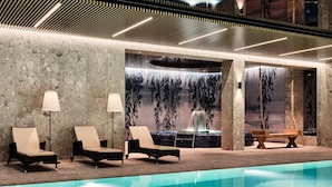 Un área de piscina interior moderna con sillas de descanso, un banco y una pared con un mural de cascada.