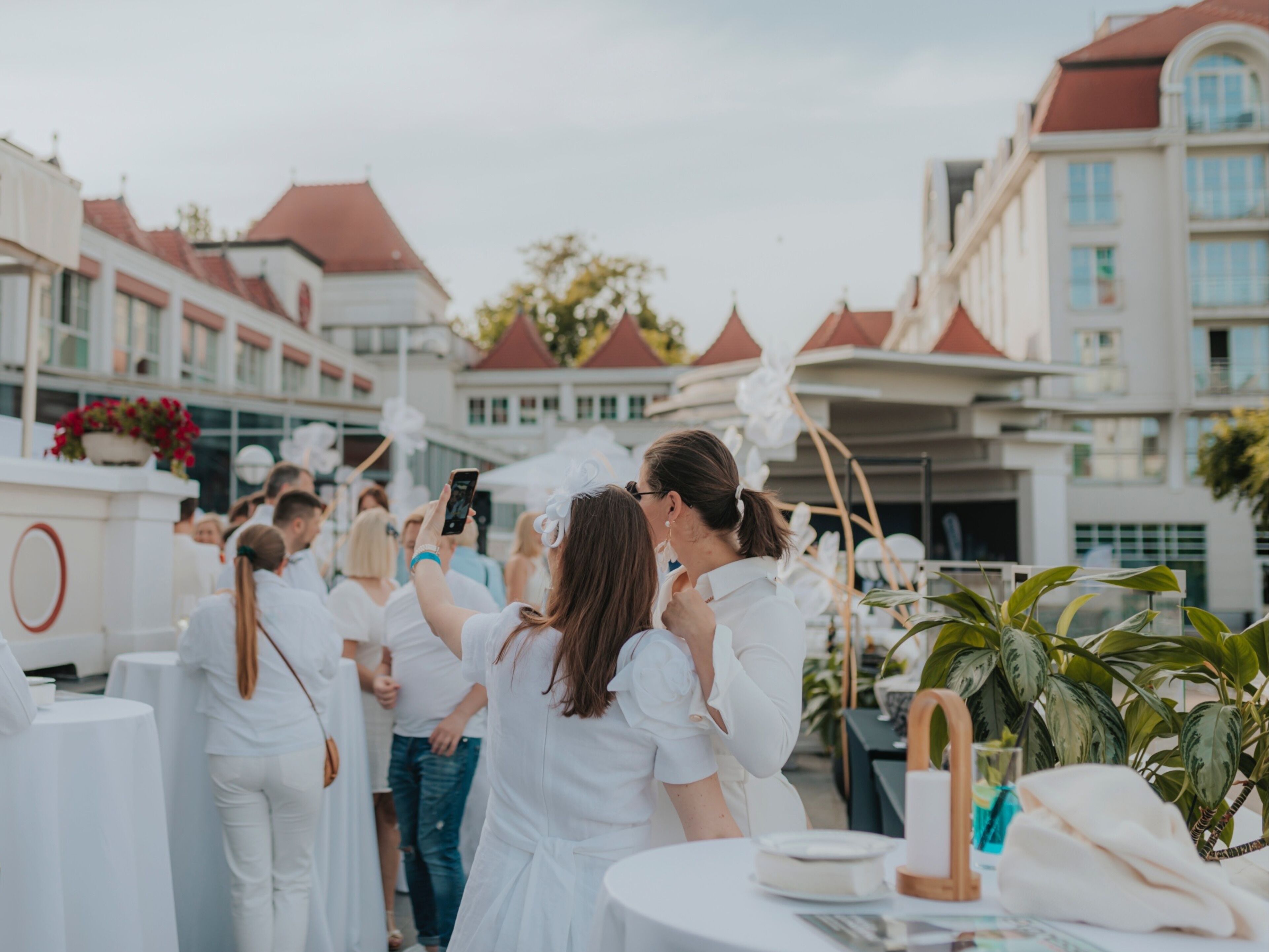 Un grupo de personas vestidas de blanco en un evento al aire libre, con un edificio de fondo que tiene techos rojos y paredes blancas.