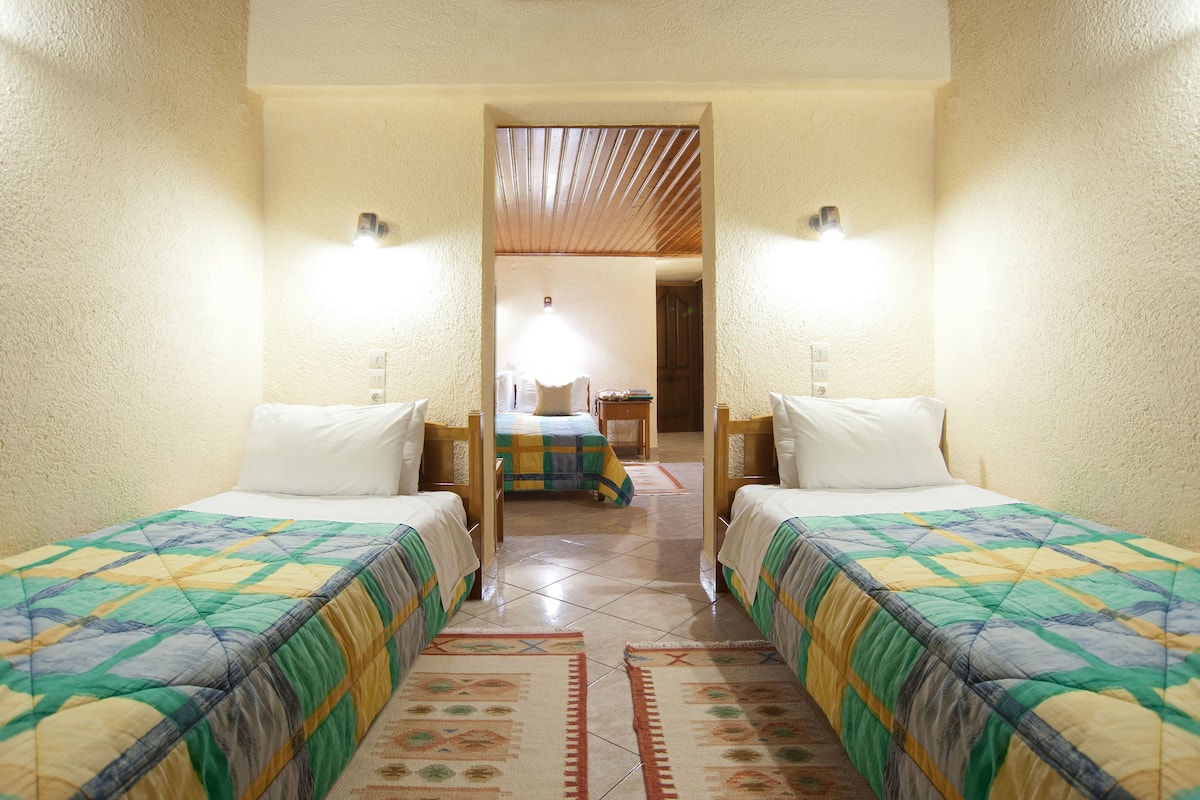 A room with two beds, a wooden ceiling, and a view into another room with a bed and a table.