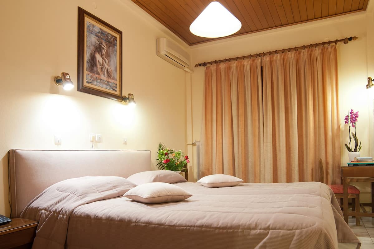 A bedroom with a large bed, a wooden ceiling, a nightstand, a desk with a chair, and a window with curtains.