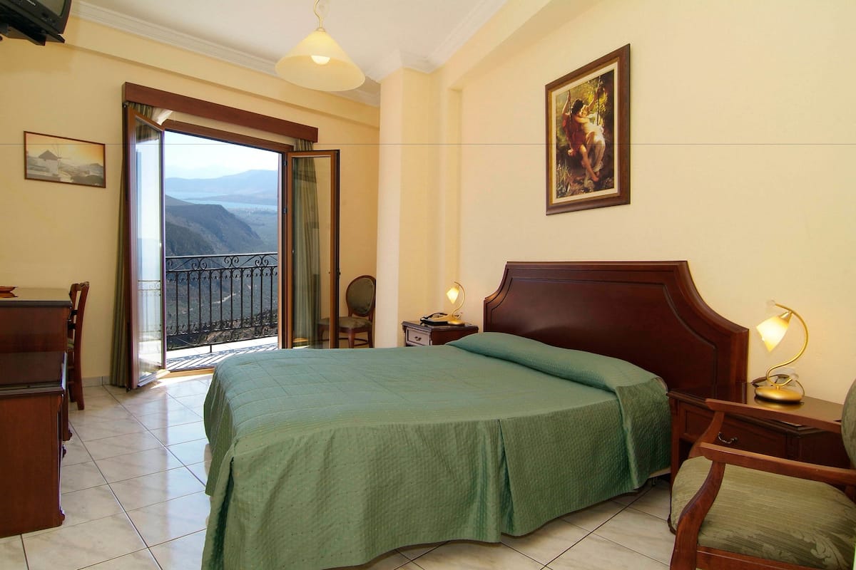 A hotel room with a large bed, a desk, a chair, a nightstand with a lamp, and a balcony with a view.