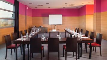 Un salón de conferencias con una larga mesa, sillas, una pantalla de proyector y un ventanal con cortinas.