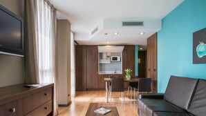 A modern living room with a sofa, a coffee table, and a TV on a wooden cabinet. There is a kitchen area with a microwave and a dining table with chairs. - Hotel Eurostars Rey Fernando (Zaragoza)