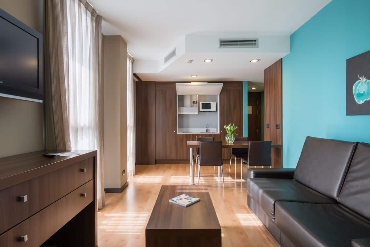 A modern living room with a sofa, a coffee table, and a TV on a wooden cabinet. There is a kitchen area with a microwave and a dining table with chairs.