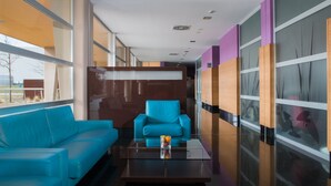 A modern hotel lobby with a blue sofa, a glass coffee table with a drink, and a large window offering a view of the outdoors. - Hotel Eurostars Rey Fernando (Zaragoza)