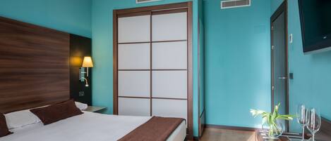 Habitación de hotel con una cama grande, cabecera de madera, mesita de noche con lámpara, puerta corredera, televisor montado en la pared y una pequeña mesa con un florero y vasos.