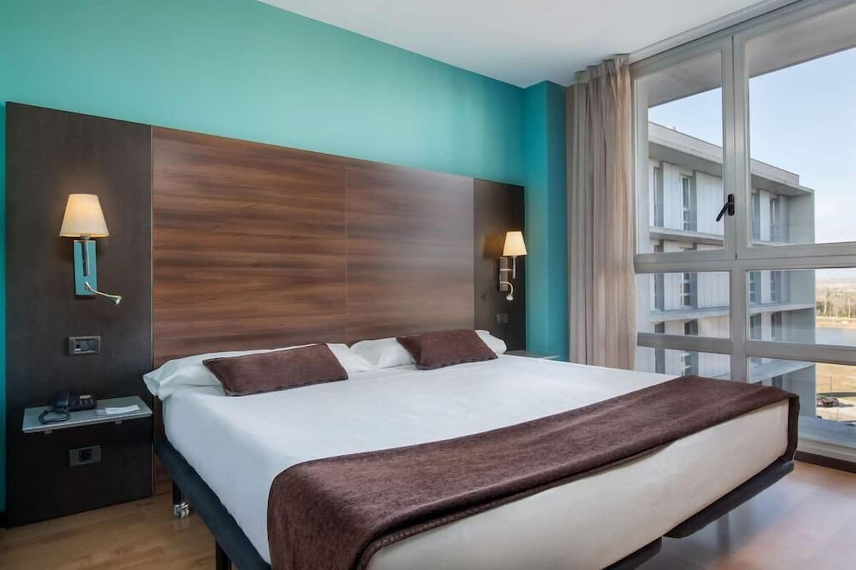 A hotel room with a large bed, two bedside lamps, a nightstand, and a window with a view of buildings.