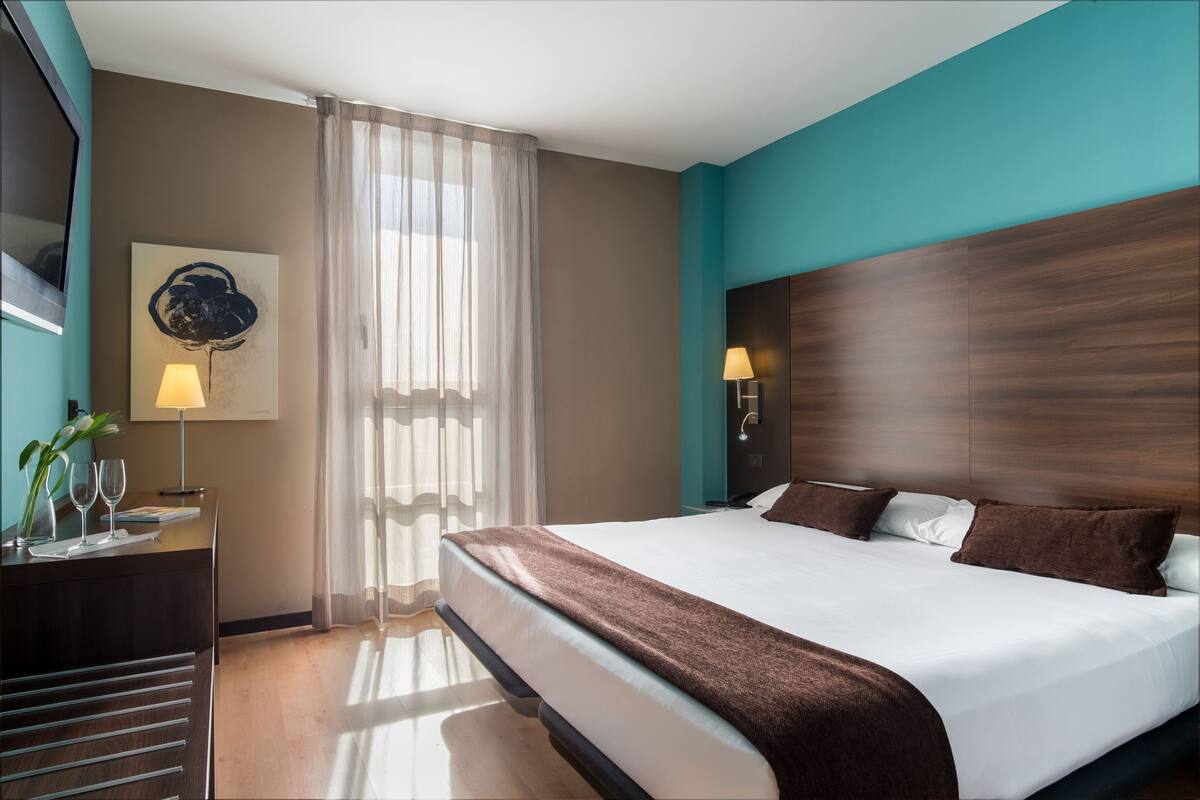 A modern hotel room with a large bed, a desk with a lamp, a painting on the wall, and a window with sheer curtains.