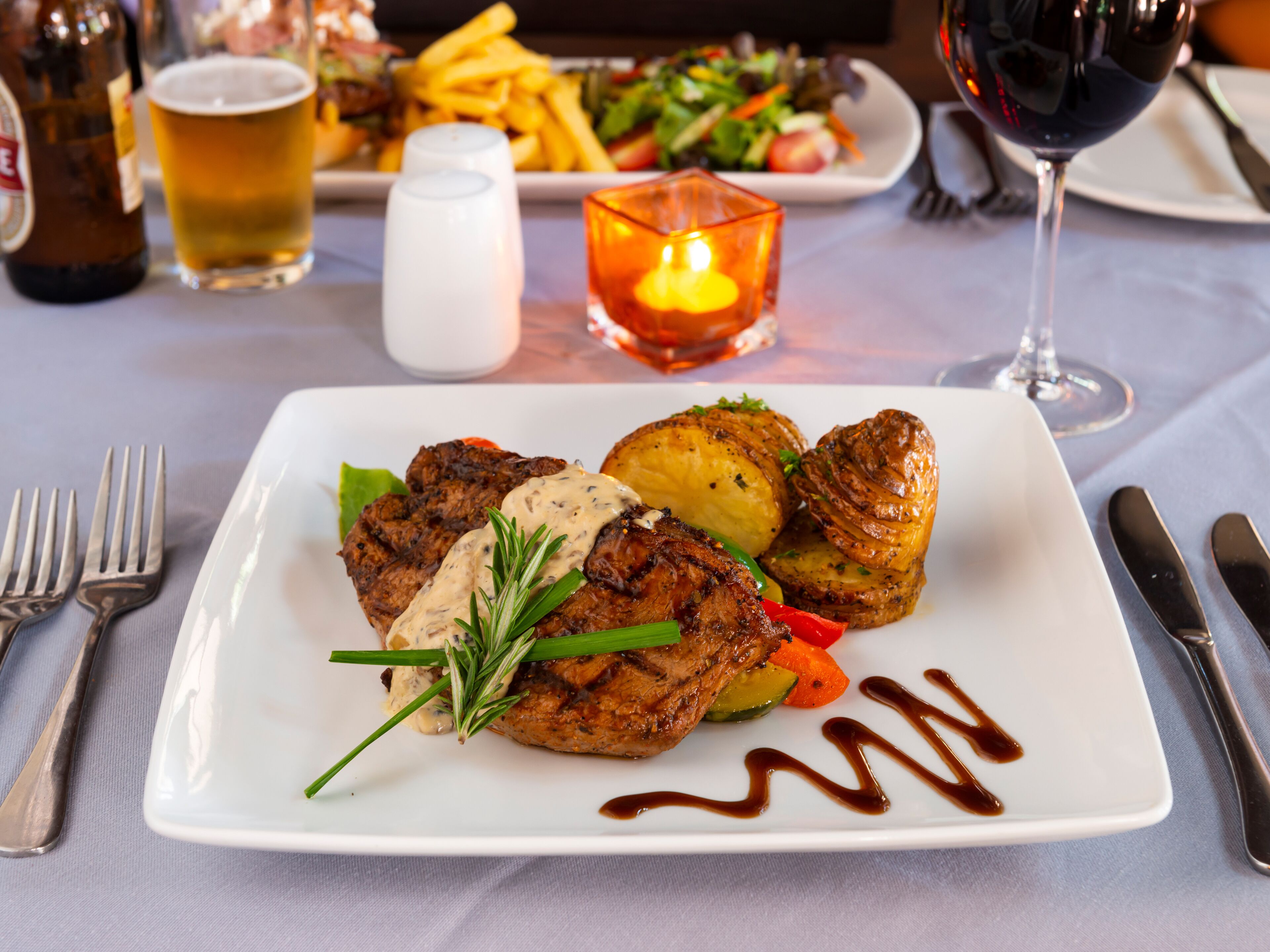A plate of food with steak, potatoes, and vegetables, a glass of red wine, and a candle on the table.