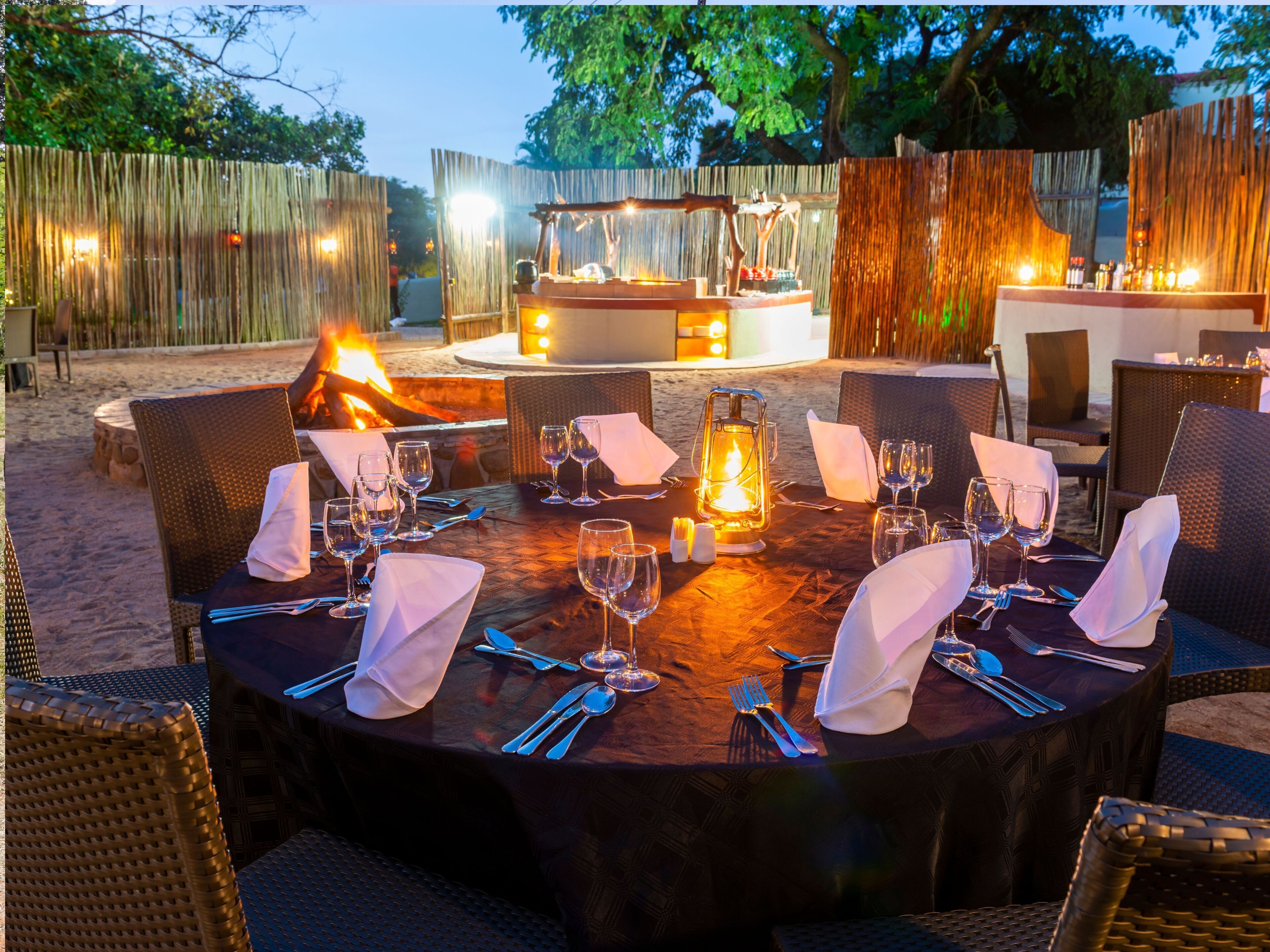 A round outdoor dining table set for a meal with a fire pit in the background.