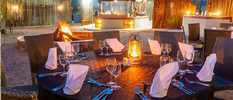 A round outdoor dining table set for a meal with a fire pit in the background.