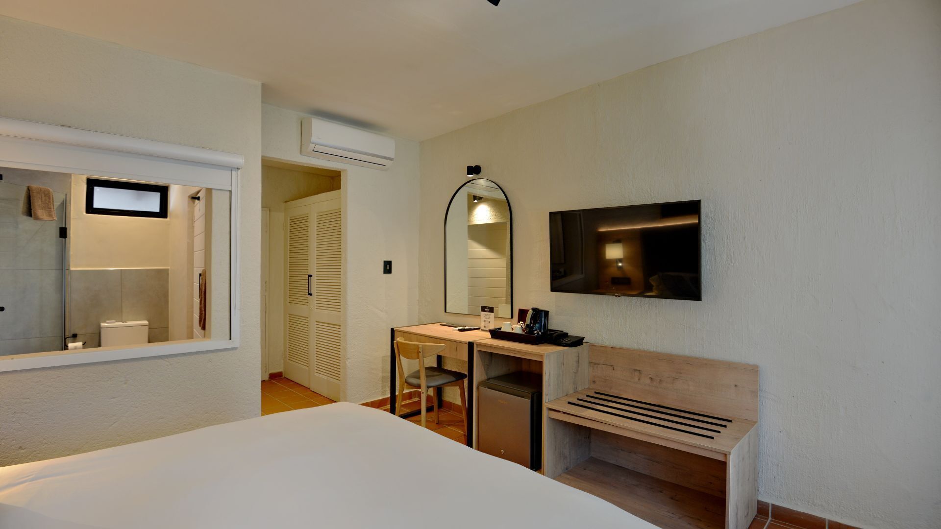 A hotel room with a bed, a desk with a chair, a mirror, a TV, and a bench.
