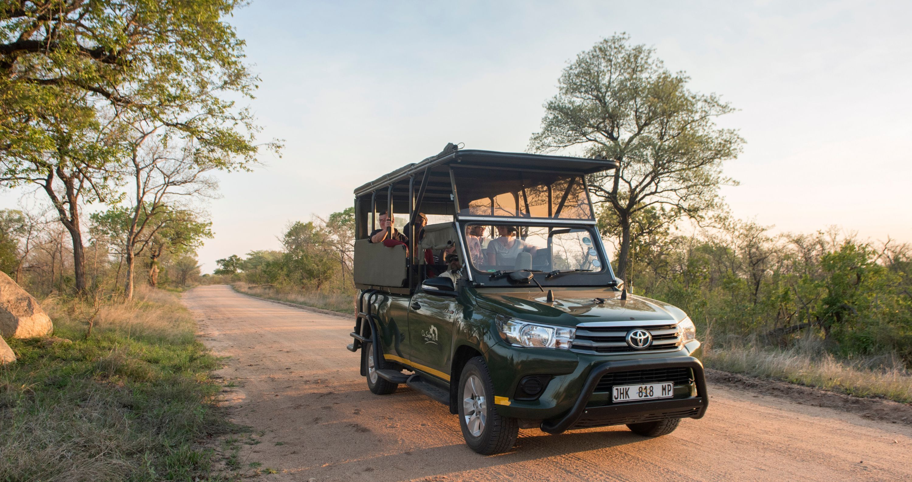 Un véhicule de safari vert, doté d’un toit et de coffres latéraux, est garé sur un chemin de terre, entouré d’arbres.