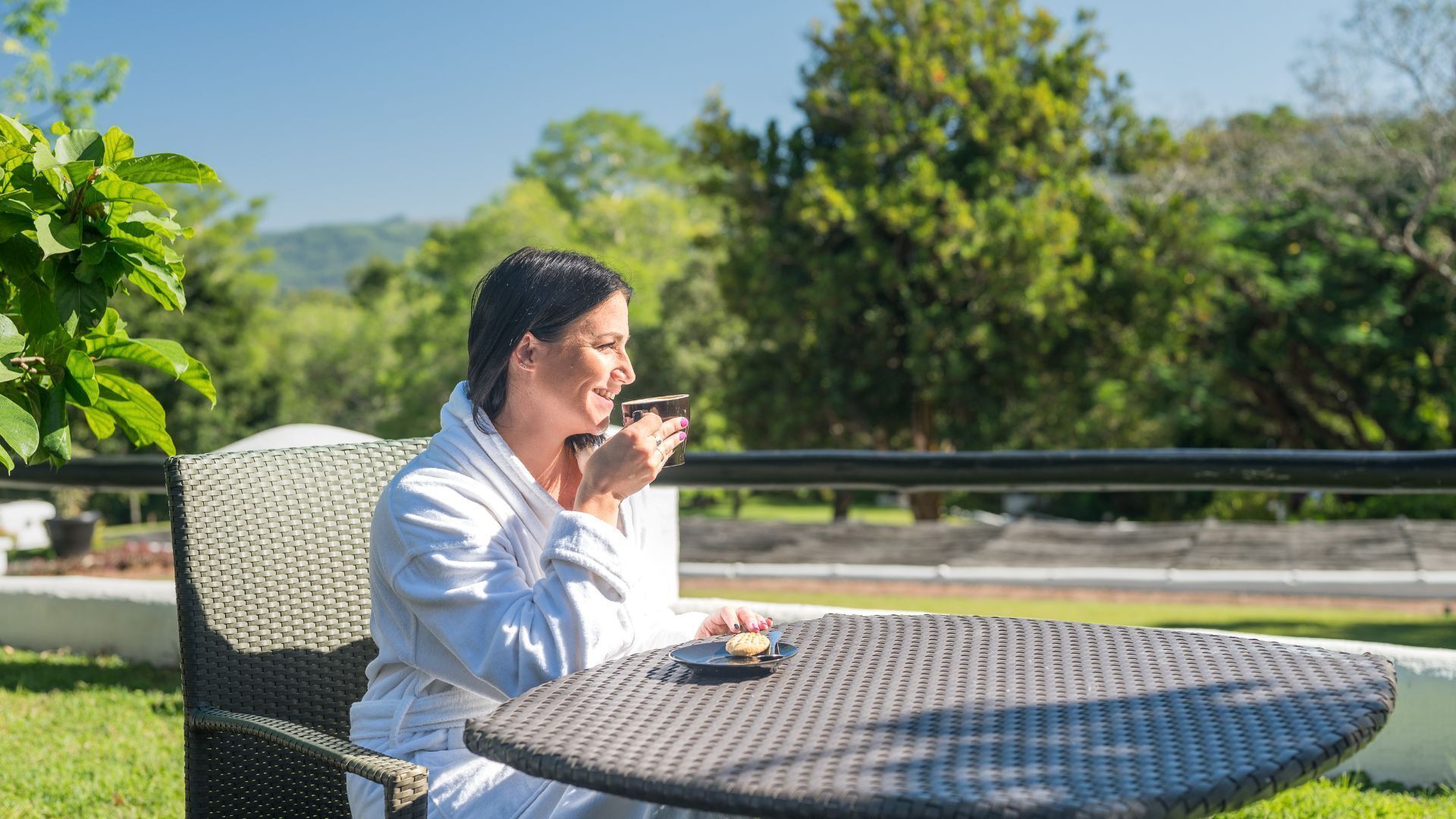 A person in a white robe sitting at an outdoor table with a cup and a cookie.