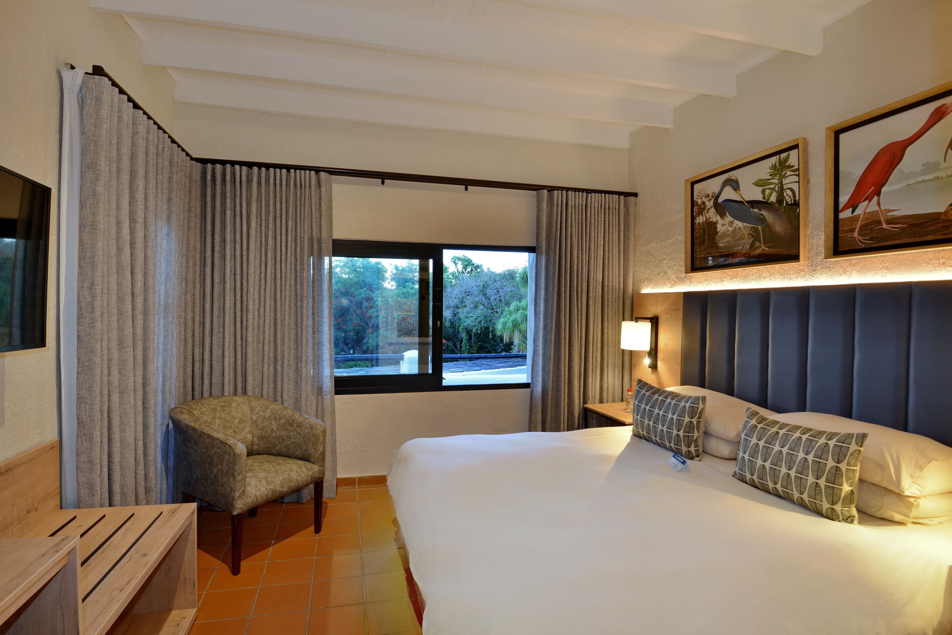 Habitación de hotel con una cama grande, una silla, un escritorio y un ventanal con cortinas.