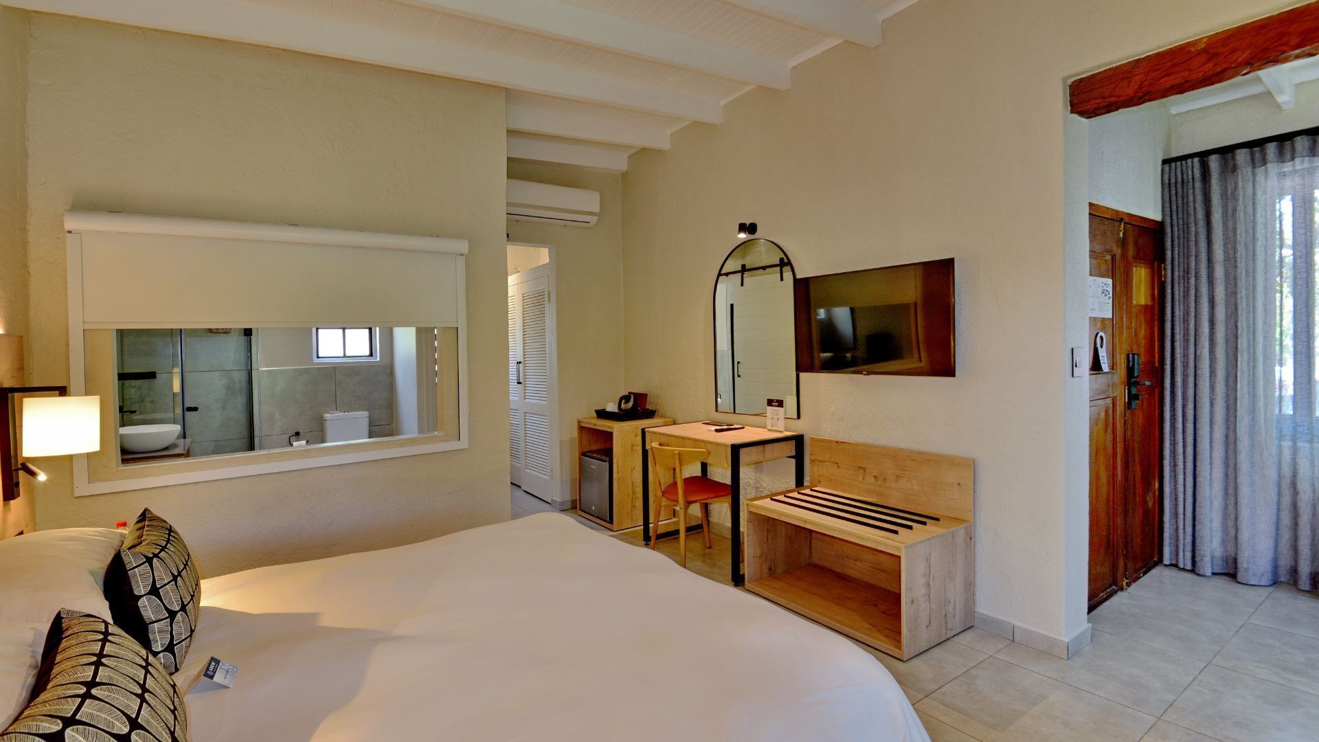 A hotel room with a large bed, a desk, a chair, a small table, a mirror, and a door.