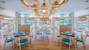 Un restaurante amplio con numerosas mesas y sillas, ventanas grandes que ofrecen vistas al exterior y un diseño de techo moderno con lámparas colgantes.