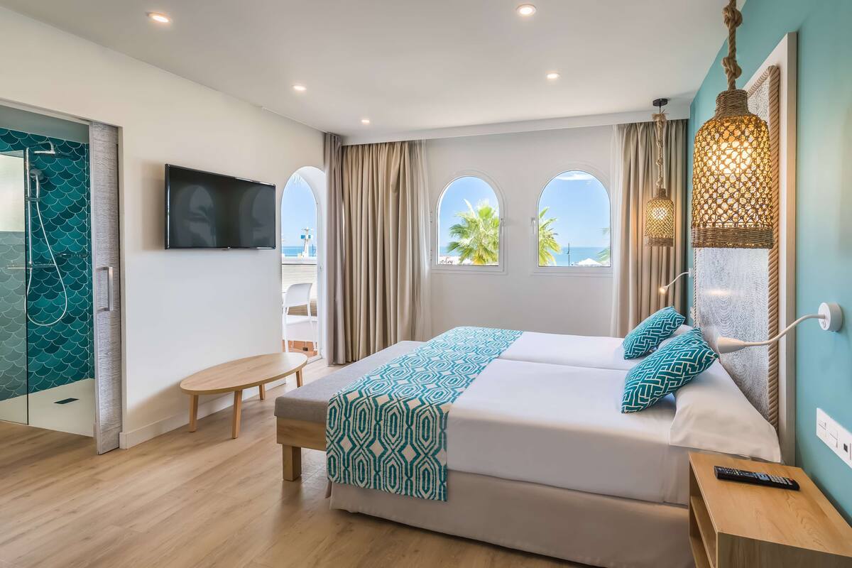 A modern hotel room with a large bed, a TV, a bench, and a view of the sea.