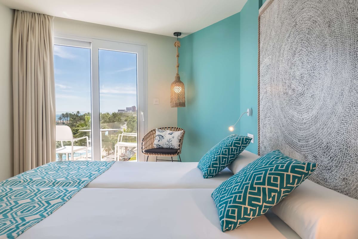 A hotel room with a large bed, two patterned pillows, a wicker chair, and a view of the outdoors.