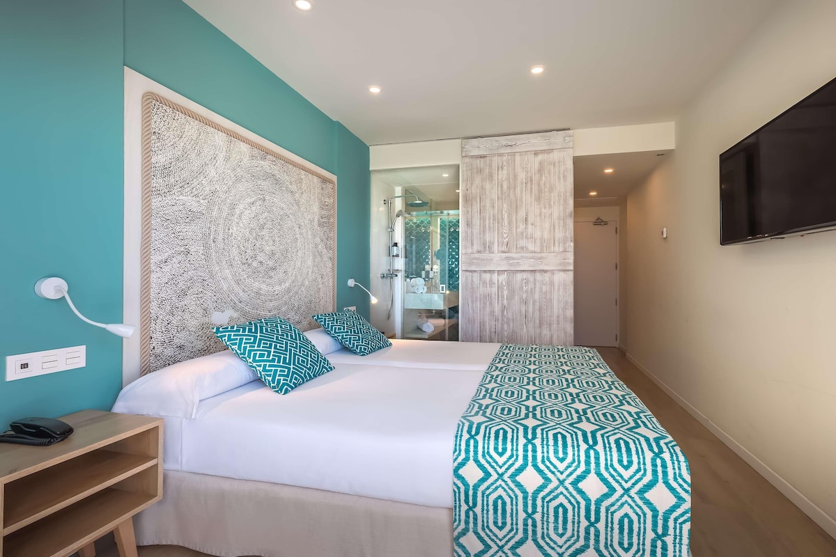 A hotel room with a bed, a bedside table, a TV mounted on the wall, and a bathroom visible through a glass door.
