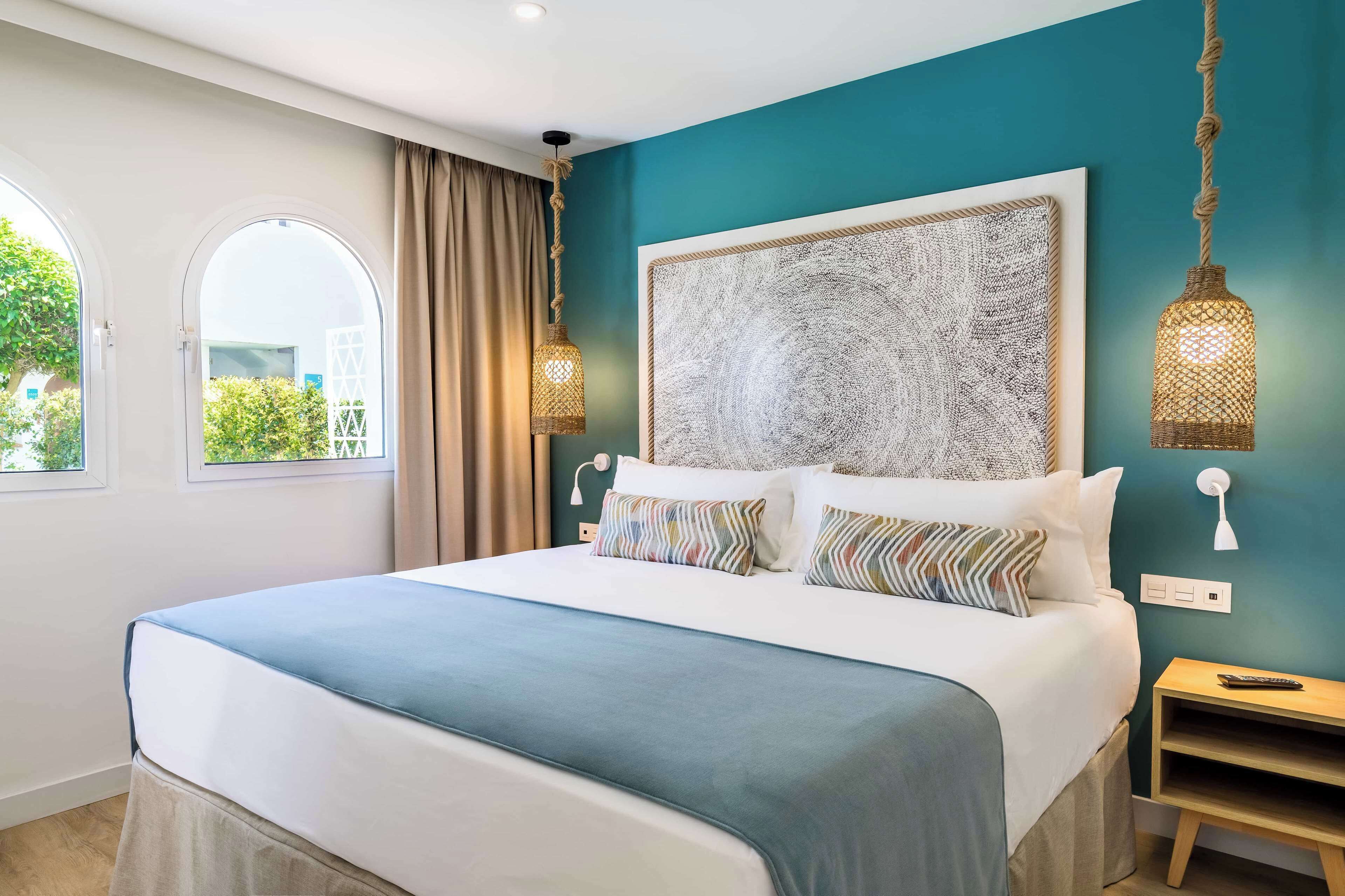 A modern bedroom with a large bed, a view of greenery outside, and a unique textured wall art piece.