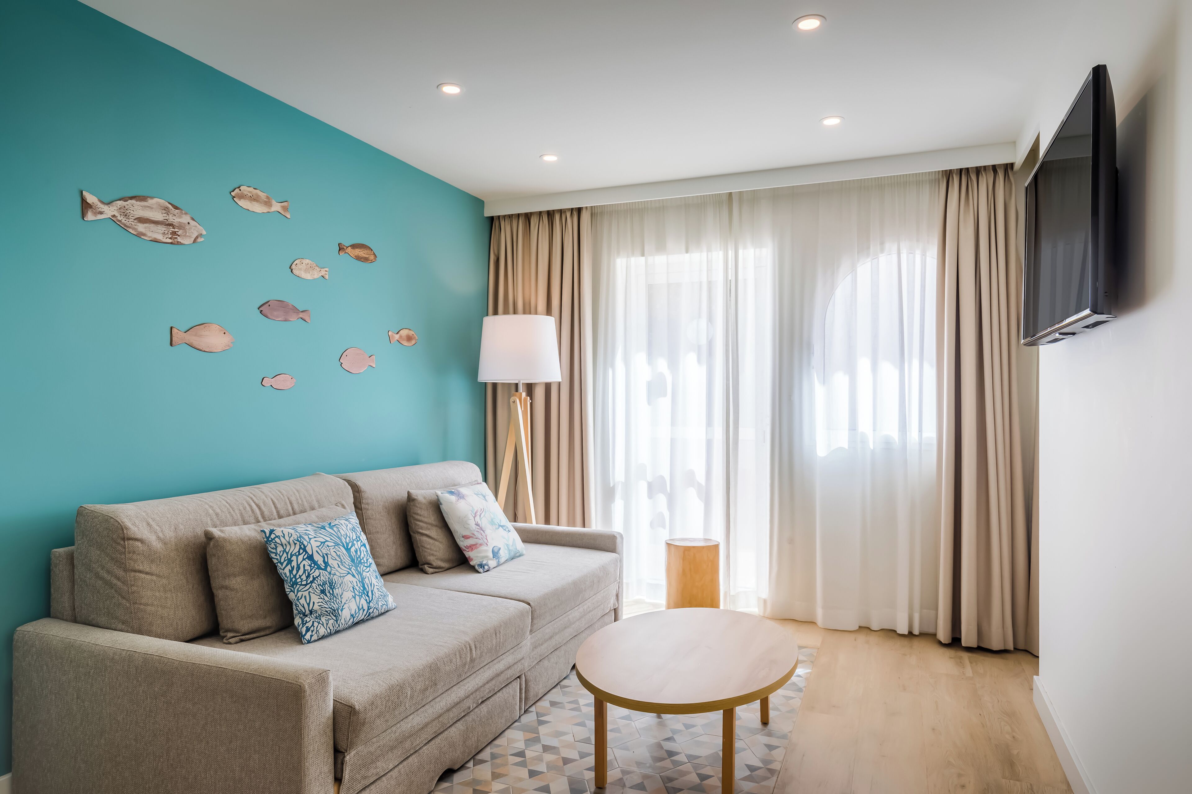 A modern living room with a teal accent wall, a beige sofa with patterned cushions, a round wooden coffee table, and a mounted TV.