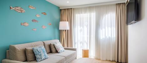 A modern living room with a teal accent wall, a beige sofa with patterned cushions, a round wooden coffee table, and a mounted TV.