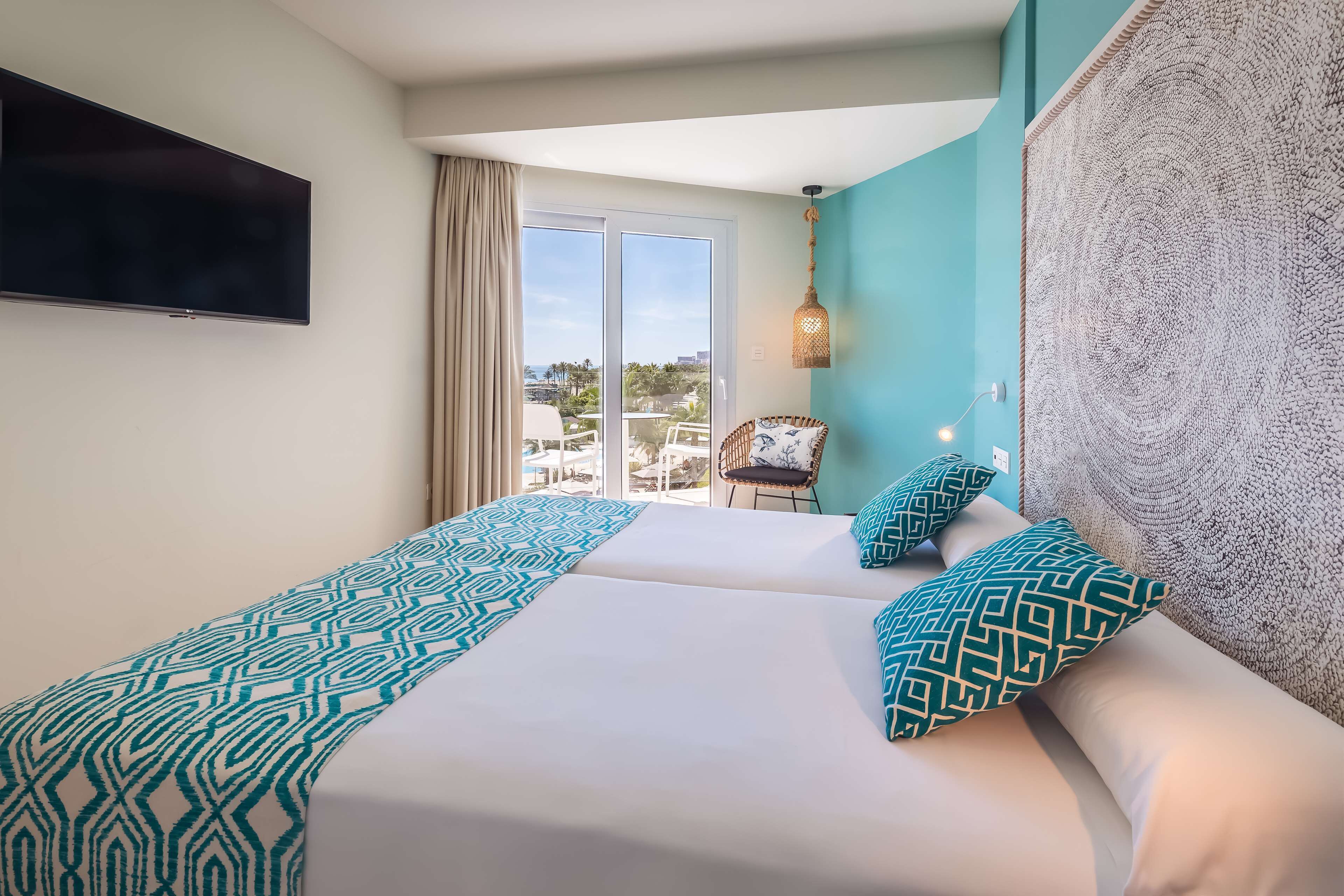 A hotel room with two beds, a TV, a balcony with a chair, and a view of the sea.