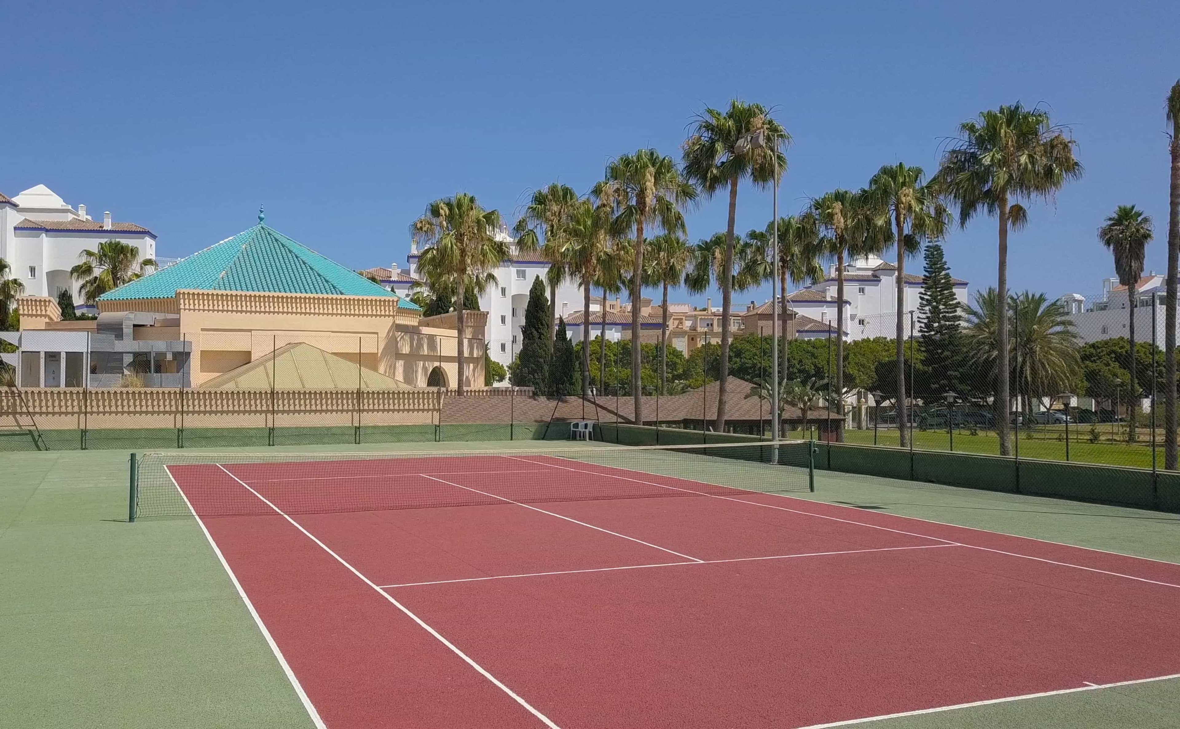 Una cancha de tenis con superficie verde y roja, rodeada por una cerca, palmeras y edificios con fachadas blancas y azules.