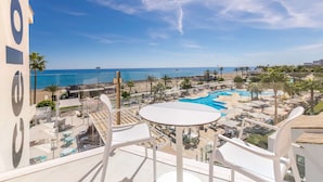 Un balcón con sillas y una mesa blancos, con vistas a una zona de resort con piscina y playa.