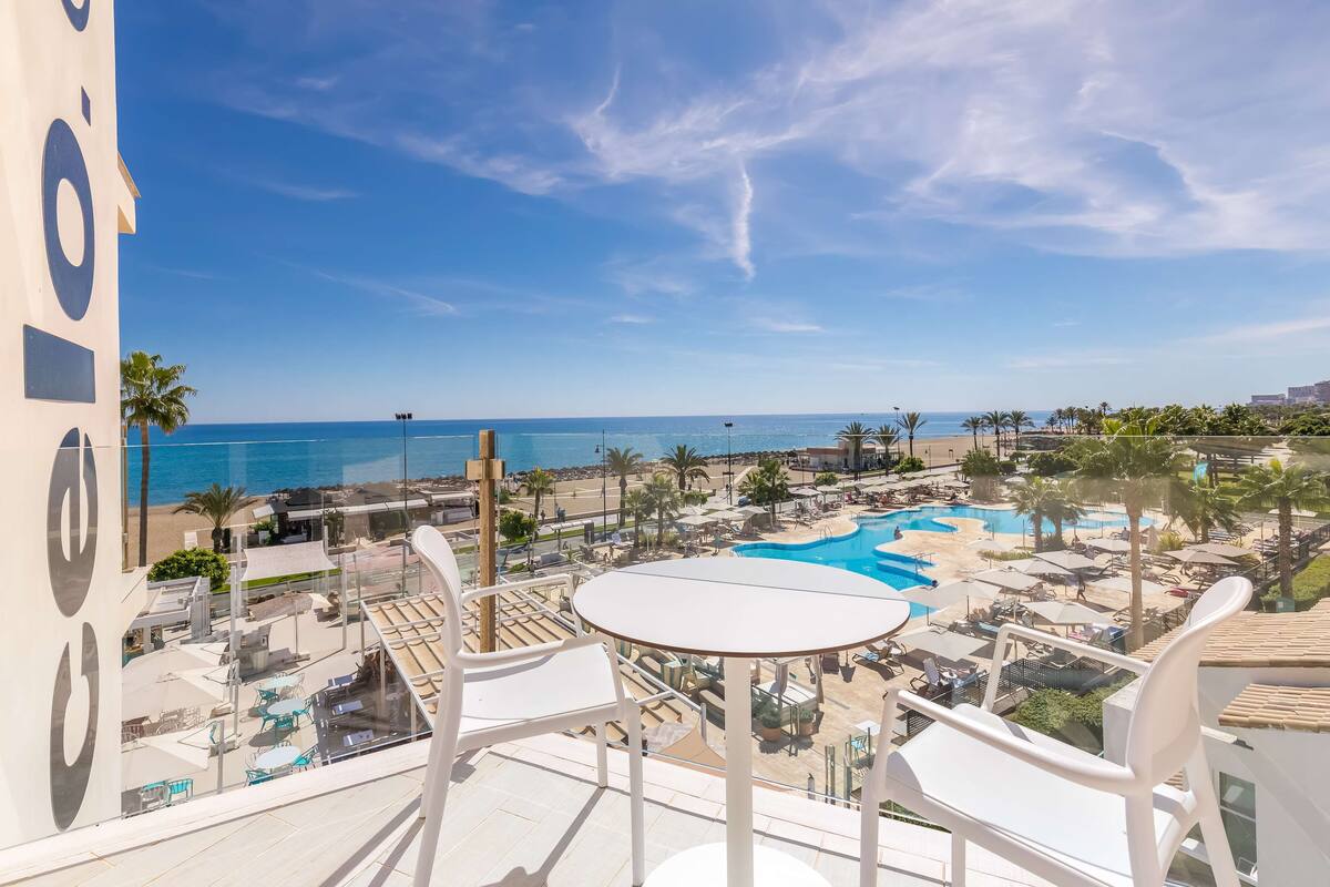 A balcony with white chairs and a table overlooking a resort area with a pool and beach.