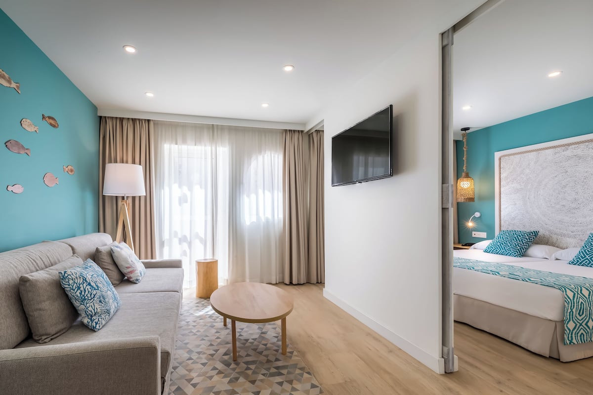 A modern hotel room with a sofa, a round table, a bed with a patterned bedspread, a flat-screen TV, and a large window with curtains.