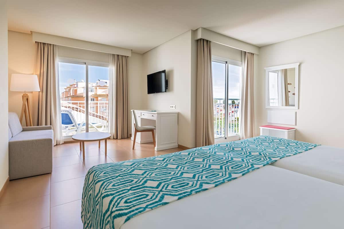 A hotel room with a bed, a desk, a chair, a TV, and a balcony with a view of buildings.