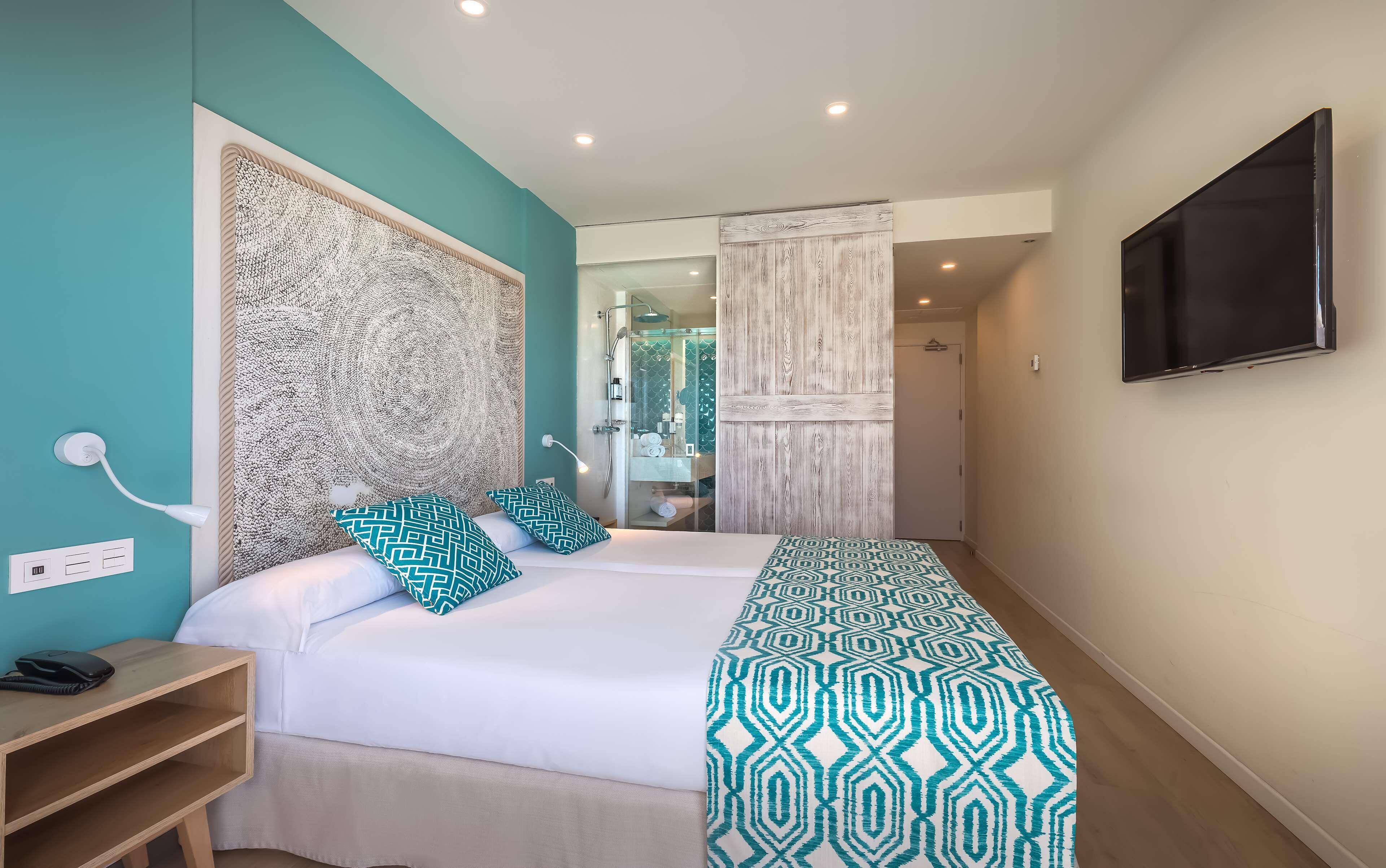 Un dormitorio con cama, mesita de noche, televisor montado en la pared y zona de ducha visible a través de un panel de vidrio.