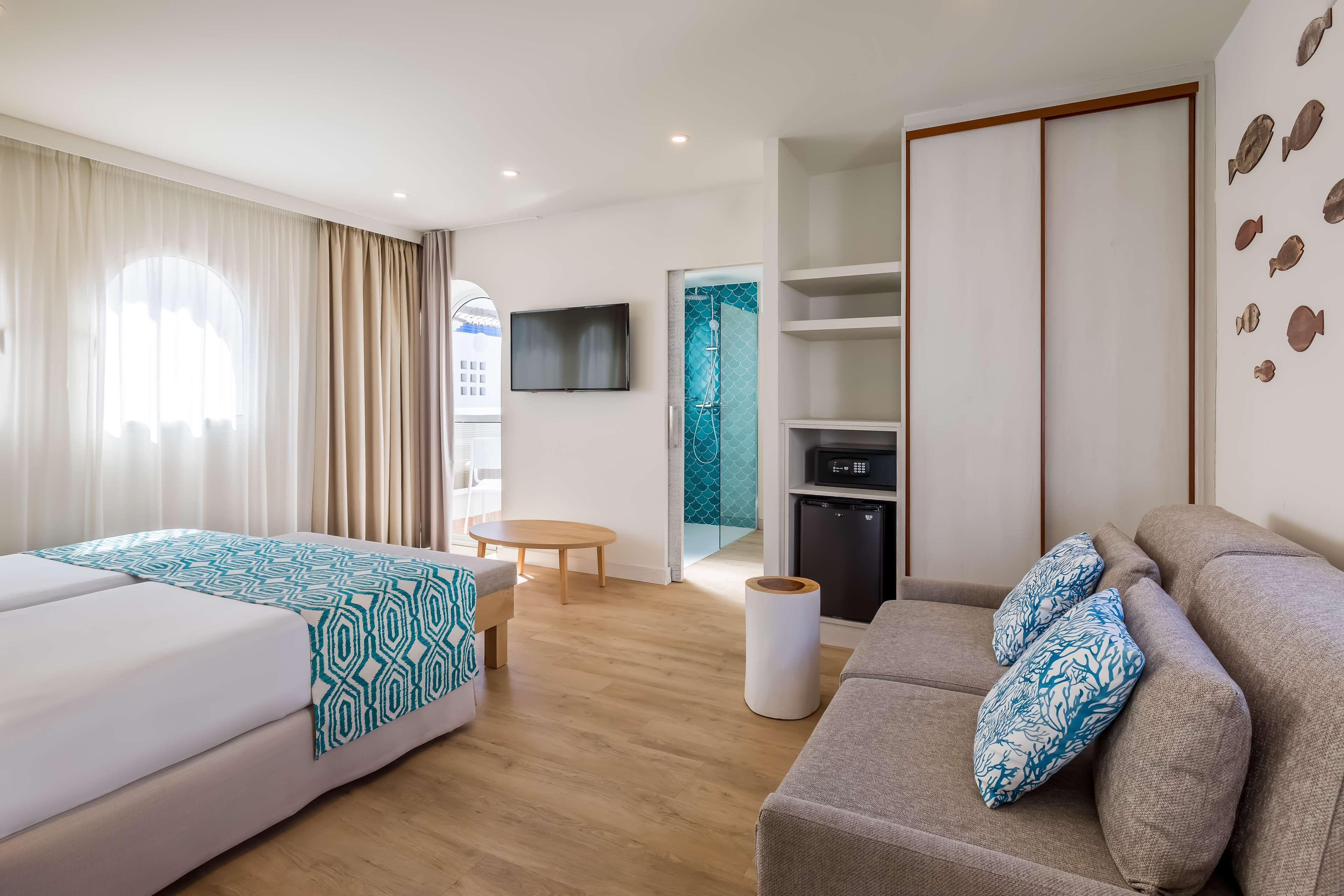 A modern hotel room with a bed, a sofa, a TV, a small table, and a bathroom with a shower.